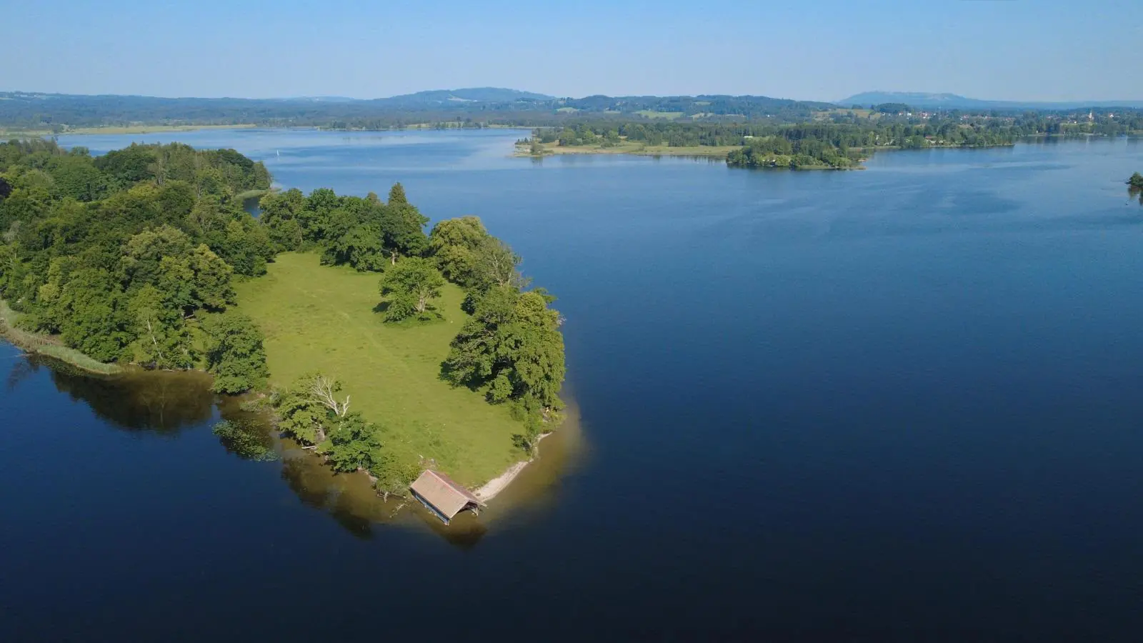 Luftaufnahme eines Sees im Voralpenland mit bewaldeter Insel und Bootshaus am Ufer.